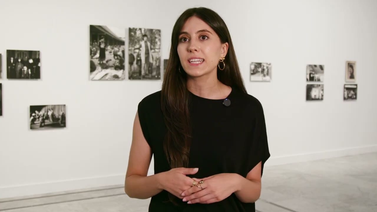 Mujer hablando ante la cámara en una sala de exposiciones, con fotografías en blanco y negro expuestas al fondo.