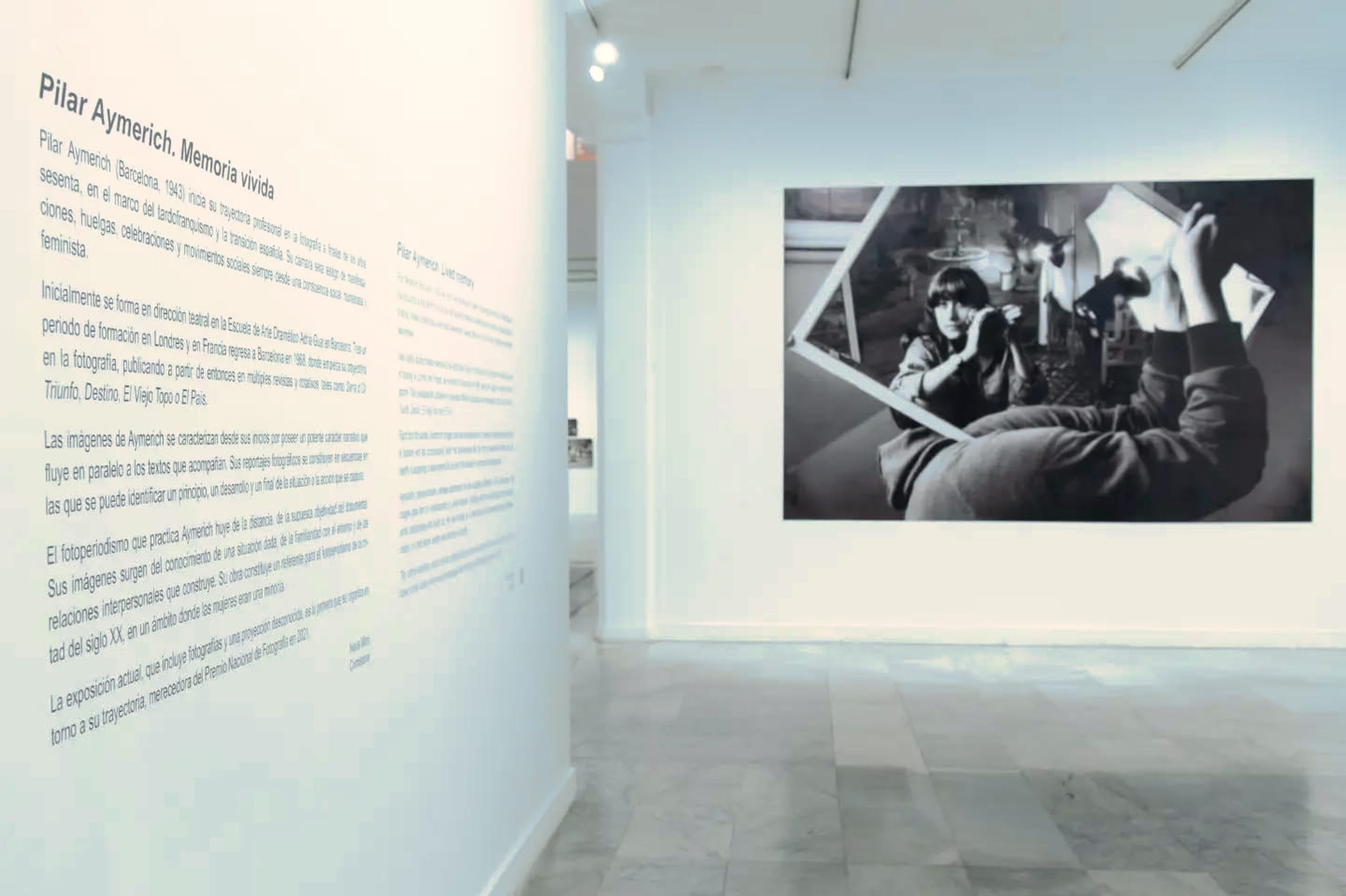 Panel de sala con el título 'Pilar Aymerich. Memoria vivida' y texto curatorial en una pared blanca; al fondo, fotografía en blanco y negro de una mujer joven recostada sosteniendo fragmentos de espejo que reflejan la escena.