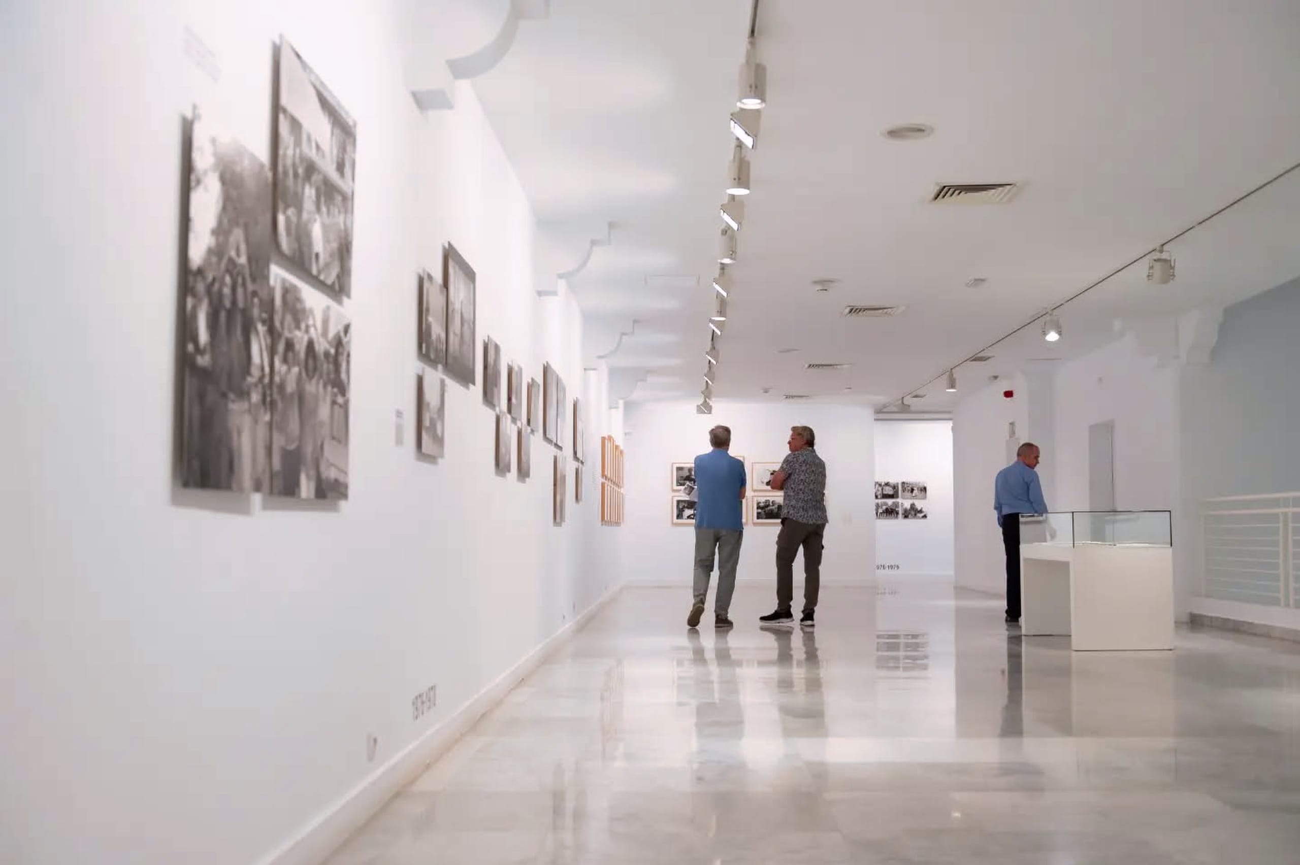Pasillo de galería con una hilera de fotografías en blanco y negro de manifestaciones callejeras; dos visitantes observan las obras y al fondo hay una vitrina expositiva.