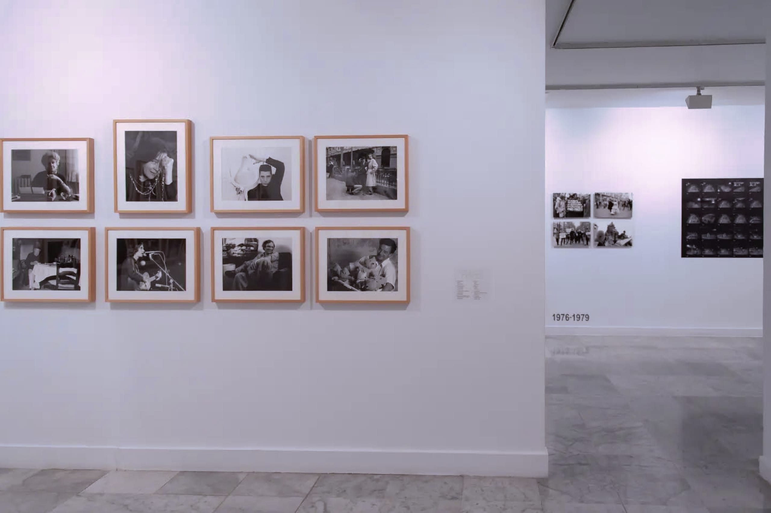 Ocho retratos en blanco y negro con marco de madera clara, colgados en dos filas de cuatro; al fondo, a través de una puerta, otra sala con fotografías pequeñas de manifestaciones y una hoja de contactos.