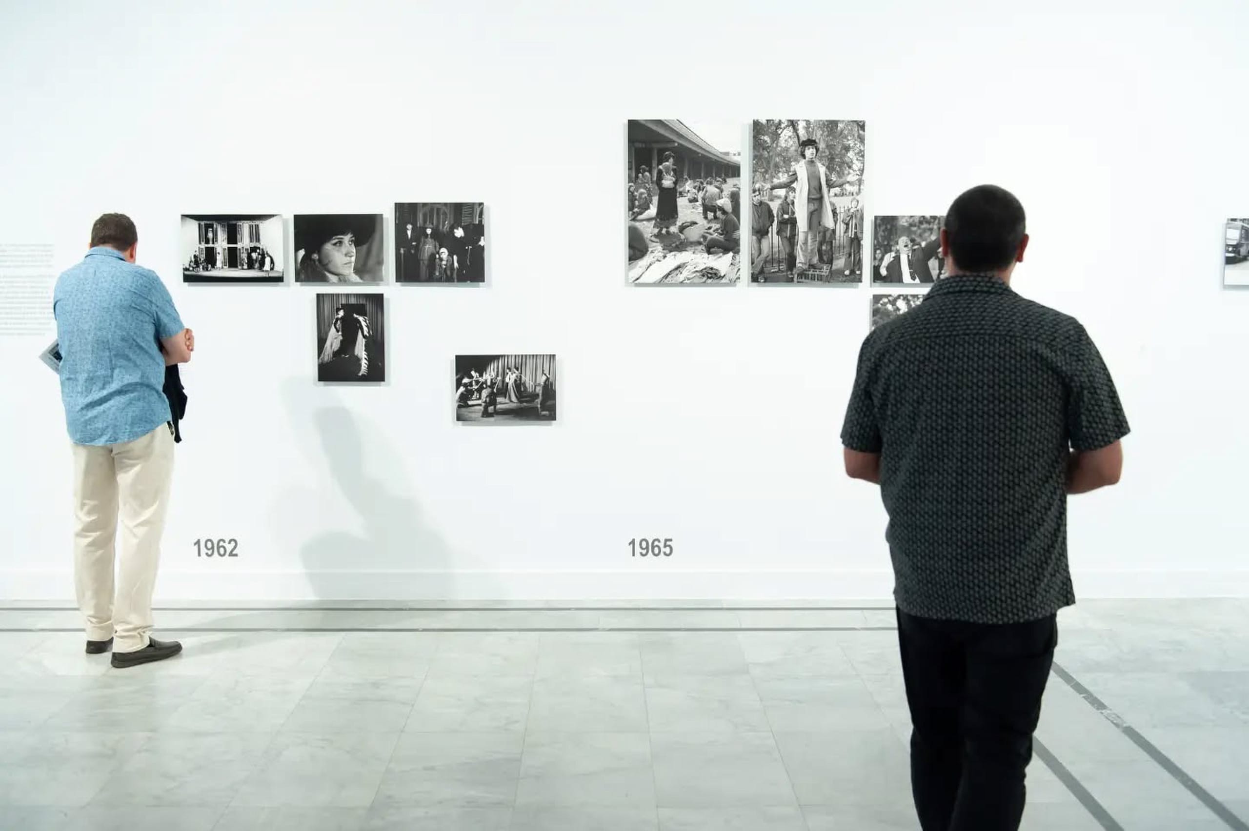 Dos visitantes de espaldas observan fotografías en blanco y negro agrupadas bajo los rótulos '1962' y '1965'; el primer grupo muestra escenas teatrales, el segundo una manifestación callejera en Londres con una persona subida a una valla.