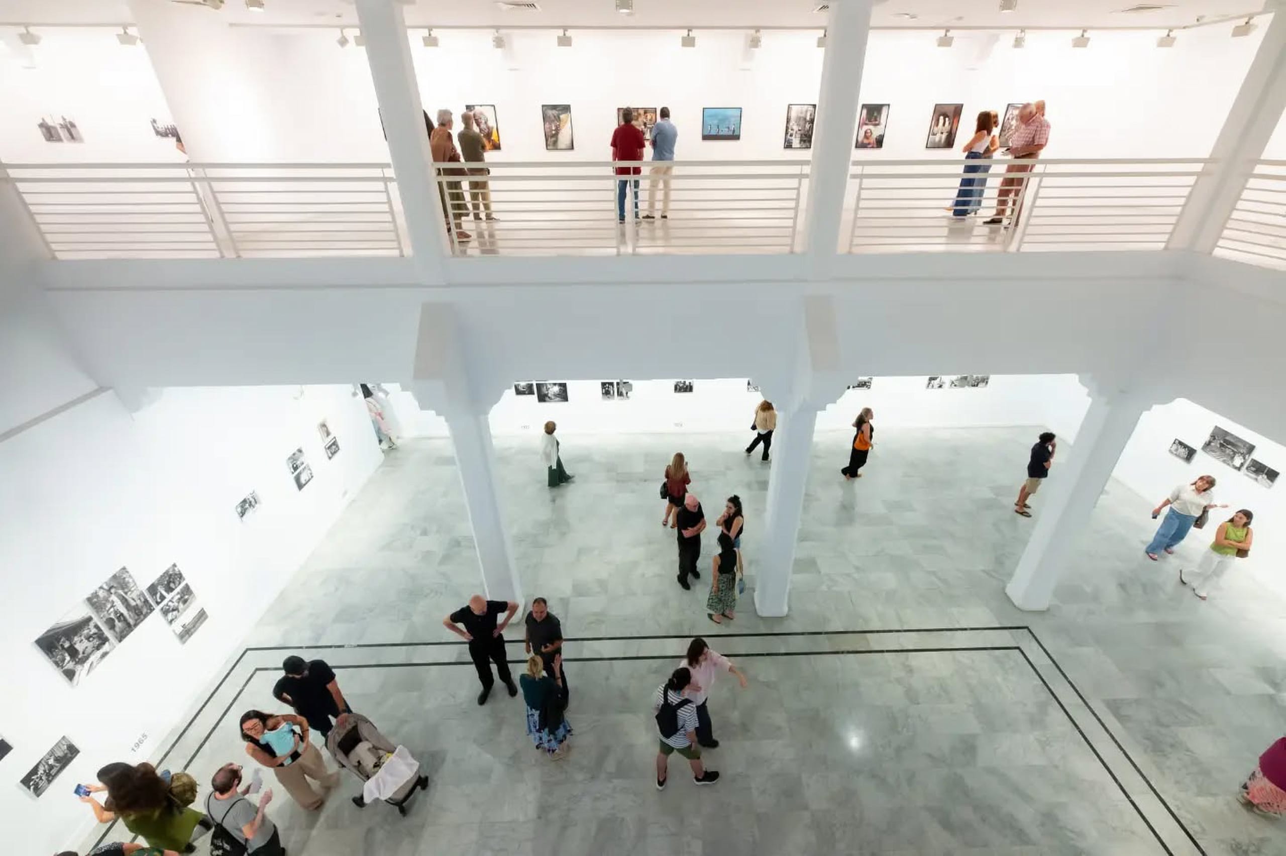 Vista desde la planta superior de una sala de exposiciones de dos plantas, con paredes blancas cubiertas de fotografías enmarcadas y visitantes en ambos niveles.