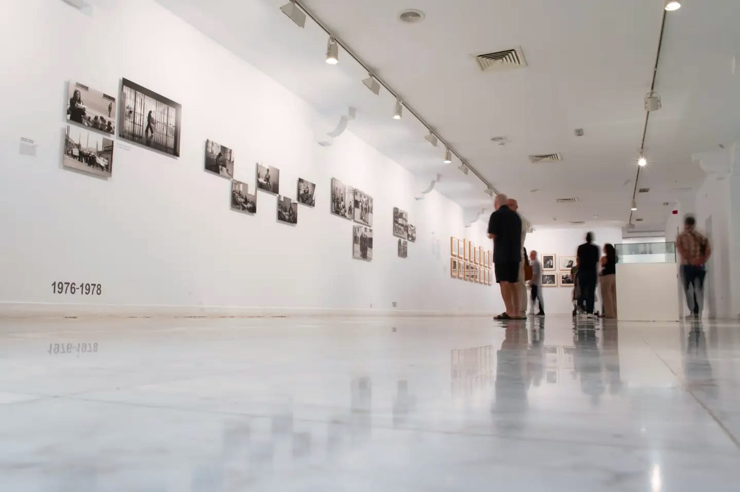 Vista de una sala de exposiciones con fotografías en blanco y negro organizadas en series. En el suelo se lee '1976-1978'; varias personas recorren la sala y una vitrina aparece al fondo.