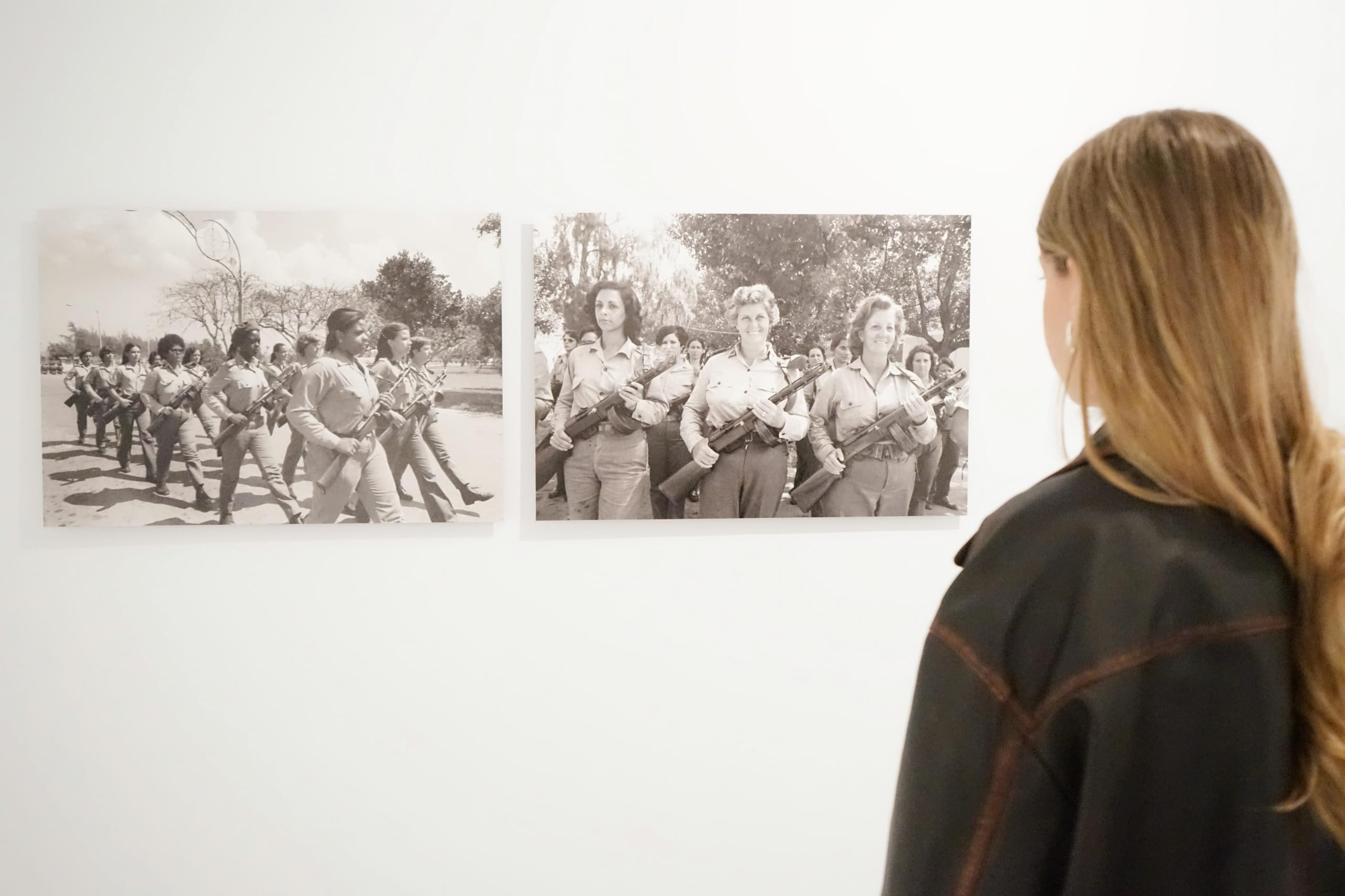 Dos fotografías en blanco y negro de mujeres uniformadas con fusiles: en una, un grupo marcha en formación; en la otra, tres mujeres posan frente a la cámara. Una visitante observa las obras de espaldas.